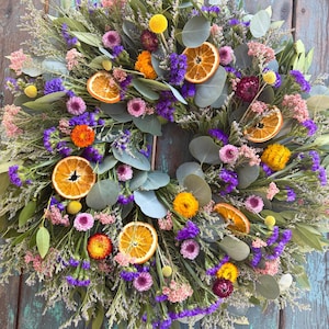 May include: A vibrant wreath featuring a variety of dried flowers and greenery. The wreath includes orange slices, purple statice, pink and yellow blooms, and eucalyptus leaves. The wreath is displayed against a weathered, turquoise-painted wooden background.