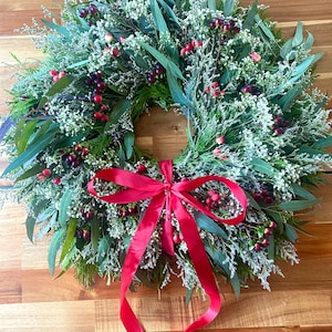 May include: A circular wreath constructed from green eucalyptus leaves, evergreen sprigs, and white filler flowers, accented with clusters of small dark purple and bright red berries. A vibrant red satin ribbon is tied in a bow at the bottom.