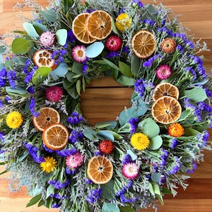 May include: A circular wreath featuring dried orange slices, vibrant purple statice flowers, green eucalyptus leaves, and small yellow, pink, and orange strawflowers. The arrangement is densely packed with various botanical elements.