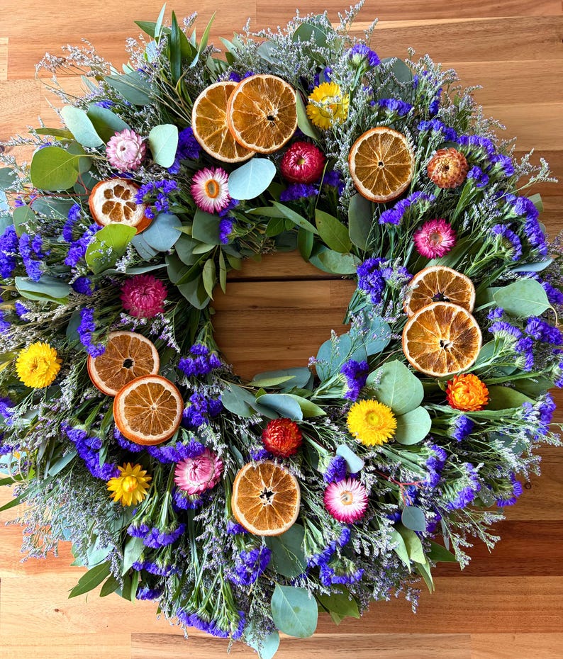 May include: A circular wreath featuring dried orange slices, vibrant purple statice flowers, green eucalyptus leaves, and small yellow, pink, and orange strawflowers. The arrangement is densely packed with various botanical elements.