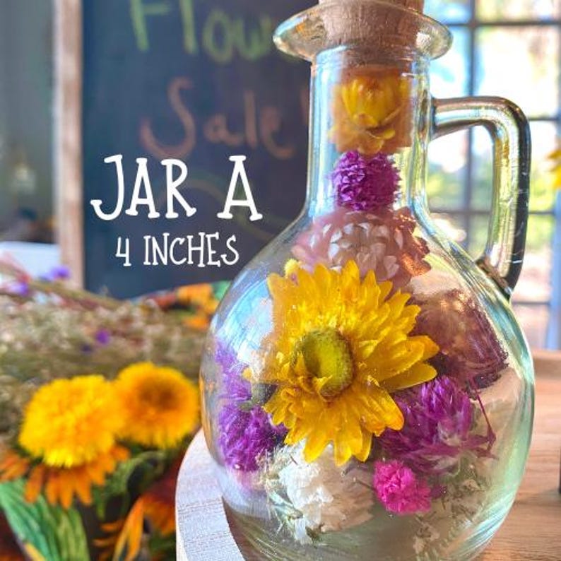 May include: A clear glass jar, approximately 10 cm tall, filled with dried yellow, purple, and white flowers. The jar has a cork stopper and a handle. Text on the image reads "Jar A" and "4 inches".