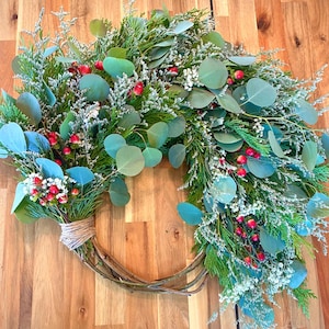 Op de afbeelding: Een handgemaakte krans met een basis van twijgen, versierd met groen gebladerte, eucalyptusbladeren, kleine witte bloemen en rode bessen. De krans is vastgebonden met touw, wat een natuurlijke, seizoensgebonden uitstraling geeft, geschikt voor huisdecoratie.
