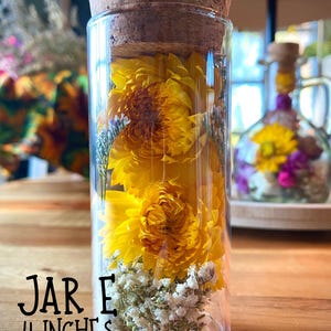 May include: A clear glass jar with a cork lid, filled with bright yellow flowers and white baby's breath. The jar is labeled "JAR E" and is 10 cm tall. The jar is sitting on a wooden surface.