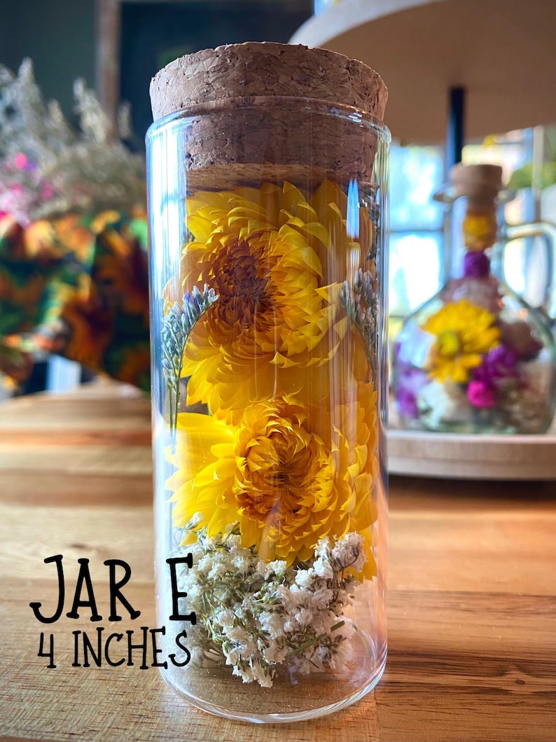 May include: A clear glass jar with a cork lid, filled with bright yellow flowers and white baby's breath. The jar is labeled "JAR E" and is 10 cm tall. The jar is sitting on a wooden surface.