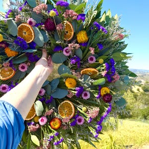 May include: A circular wreath featuring dried eucalyptus leaves, vibrant purple statice, pink flowers, yellow strawflowers, and dried orange slices. The arrangement is densely packed with various botanical elements, creating a colorful and textured display.