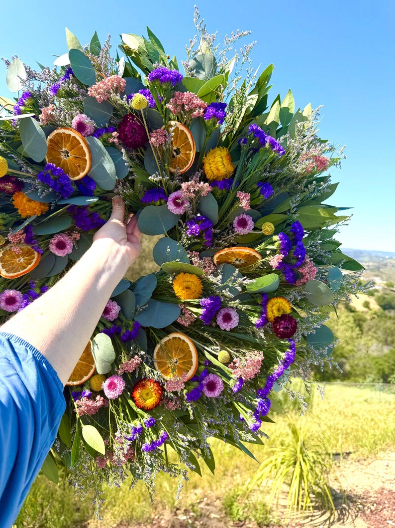 May include: A circular wreath featuring dried eucalyptus leaves, vibrant purple statice, pink flowers, yellow strawflowers, and dried orange slices. The arrangement is densely packed with various botanical elements, creating a colorful and textured display.