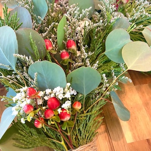 Op de afbeelding: Een boeket van groen en rode bessen, vastgebonden met touw. De compositie bevat grote, ronde, groene eucalyptusbladeren, kleine witte bloemen en groene bladeren. De bessen zijn felrood.