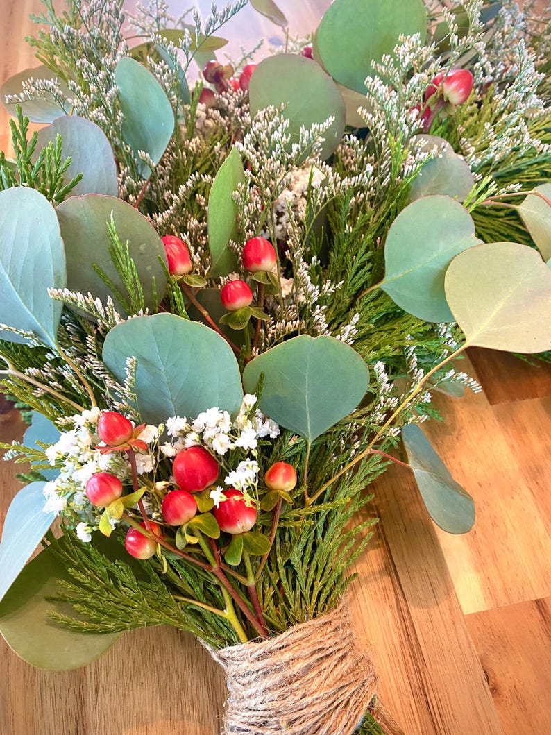 Op de afbeelding: Een boeket van groen en rode bessen, vastgebonden met touw. De compositie bevat grote, ronde, groene eucalyptusbladeren, kleine witte bloemen en groene bladeren. De bessen zijn felrood.