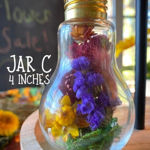 May include: A clear glass jar shaped like a lightbulb, filled with colorful dried flowers. The jar is labeled "JAR C" and is 10 cm tall. The flowers include yellow, purple, and red blooms, with green moss at the base. The jar has a gold-colored metal base.