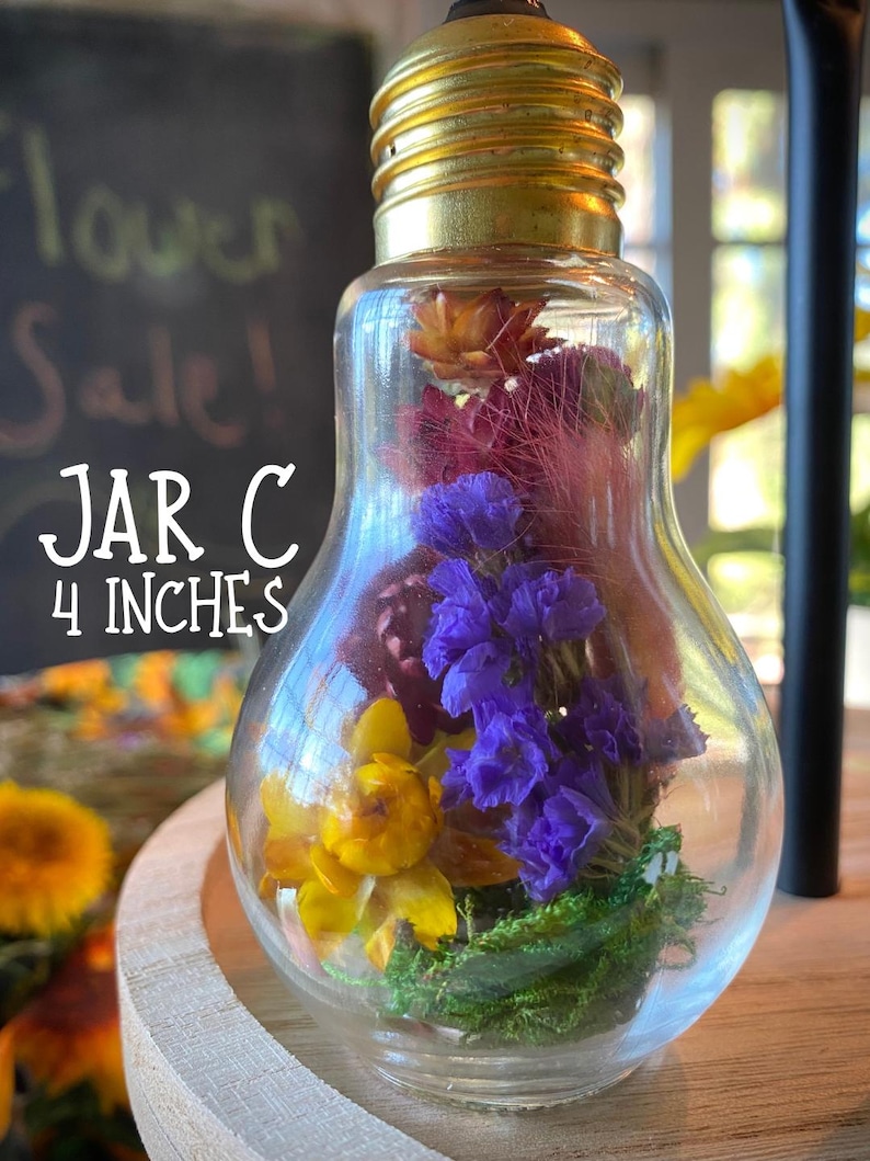 May include: A clear glass jar shaped like a lightbulb, filled with colorful dried flowers. The jar is labeled "JAR C" and is 10 cm tall. The flowers include yellow, purple, and red blooms, with green moss at the base. The jar has a gold-colored metal base.