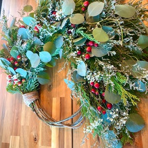 Op de afbeelding: Een handgemaakte krans met een takkenbasis, versierd met groen gebladerte, eucalyptusbladeren, kleine witte bloemen en rode bessen. De krans is vastgebonden met touw en wordt gepresenteerd tegen een houten achtergrond.