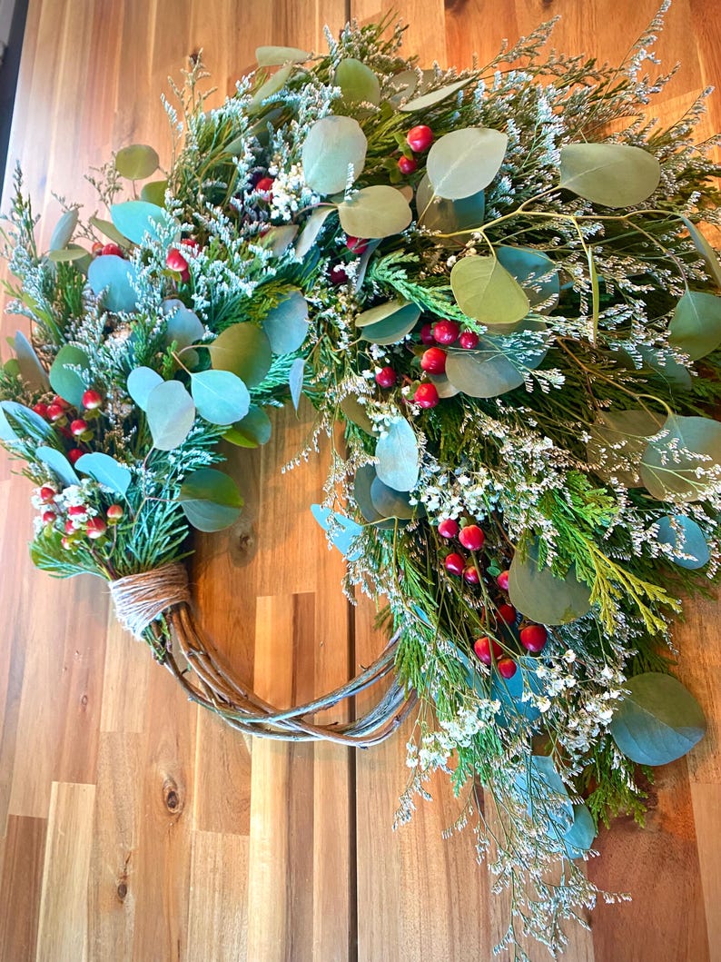Op de afbeelding: Een handgemaakte krans met een takkenbasis, versierd met groen gebladerte, eucalyptusbladeren, kleine witte bloemen en rode bessen. De krans is vastgebonden met touw en wordt gepresenteerd tegen een houten achtergrond.