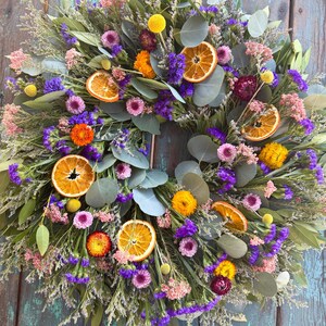 May include: A circular wreath featuring dried eucalyptus leaves, vibrant purple statice, pink filler flowers, yellow billy buttons, and dried orange slices. The arrangement is densely packed with various botanical elements, creating a textured, multi-colored display of preserved natural foliage and fruit.