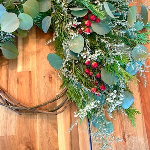 Op de afbeelding: Een handgemaakte krans met een basis van natuurlijke wijnstok, versierd met groene eucalyptusbladeren, witte bloemen en heldere rode bessen. Een feestelijke decoratie voor in huis.
