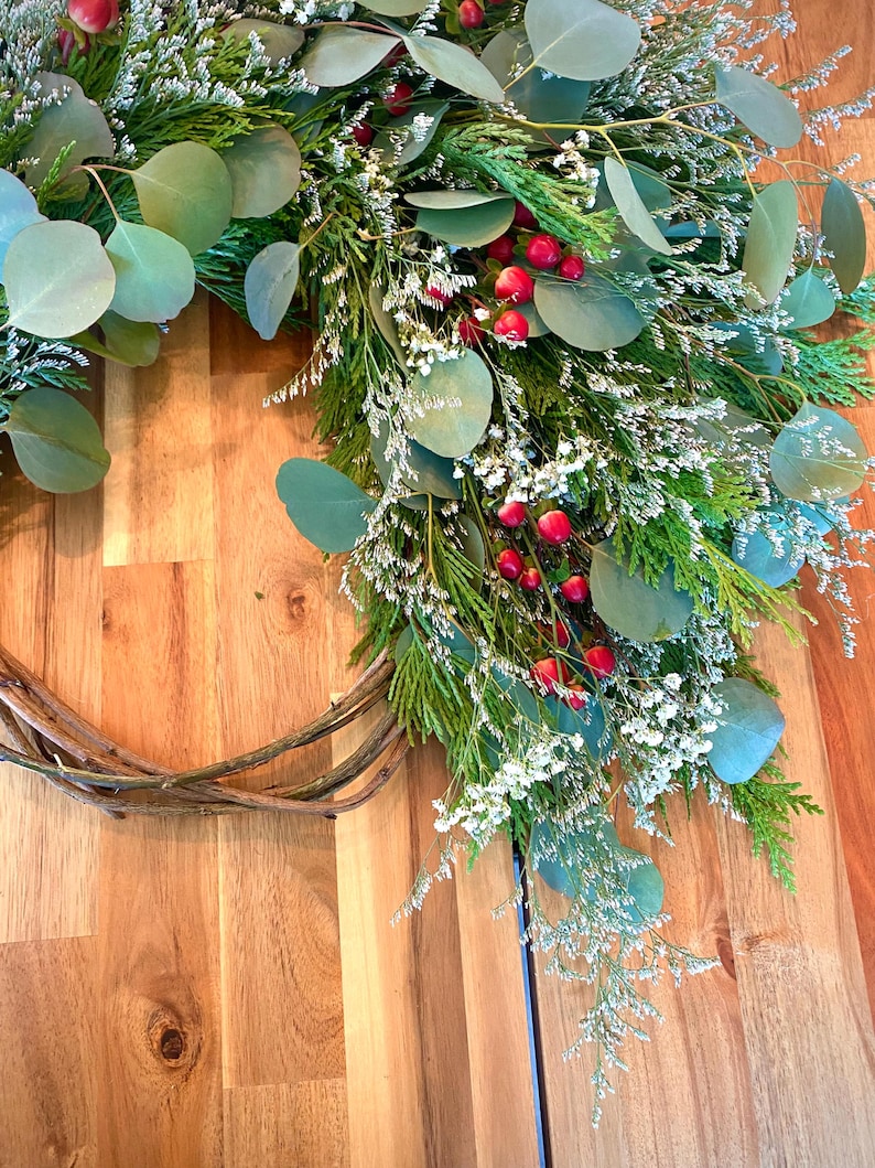 Op de afbeelding: Een handgemaakte krans met een basis van natuurlijke wijnstok, versierd met groene eucalyptusbladeren, witte bloemen en heldere rode bessen. Een feestelijke decoratie voor in huis.