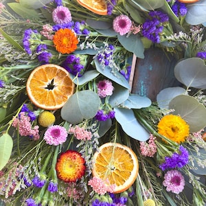 May include: A decorative wreath featuring dried orange slices, purple statice, pink strawflowers, yellow billy balls, and green eucalyptus leaves. The arrangement includes various dried botanical elements in vibrant shades of orange, purple, pink, and yellow, creating a textured, circular floral display.