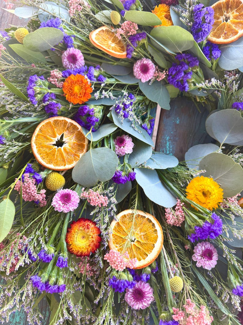 May include: A decorative wreath featuring dried orange slices, purple statice, pink strawflowers, yellow billy balls, and green eucalyptus leaves. The arrangement includes various dried botanical elements in vibrant shades of orange, purple, pink, and yellow, creating a textured, circular floral display.