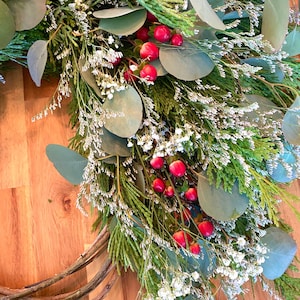 Op de afbeelding: Een handgemaakte krans met groene bladeren, eucalyptus, ceder takken en kleine witte bloemen. Rode bessen voegen kleur toe. De krans is gemaakt op een natuurlijke wijnstok basis, perfect voor seizoensgebonden decoratie.