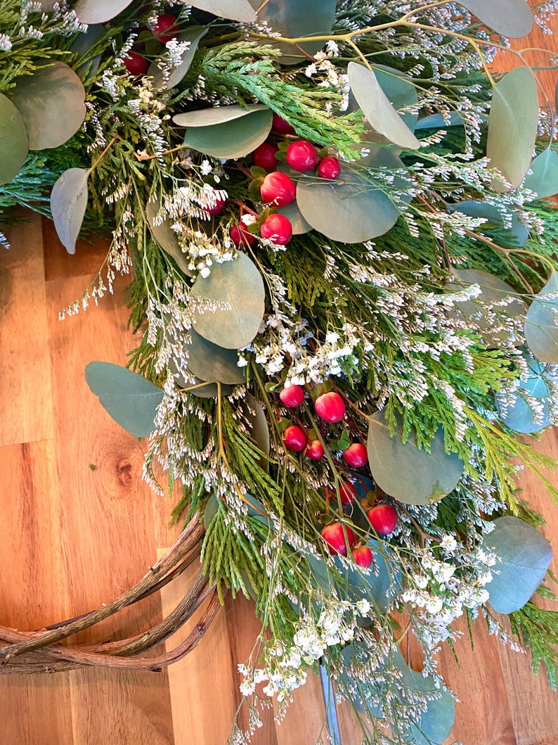 Op de afbeelding: Een handgemaakte krans met groene bladeren, eucalyptus, ceder takken en kleine witte bloemen. Rode bessen voegen kleur toe. De krans is gemaakt op een natuurlijke wijnstok basis, perfect voor seizoensgebonden decoratie.