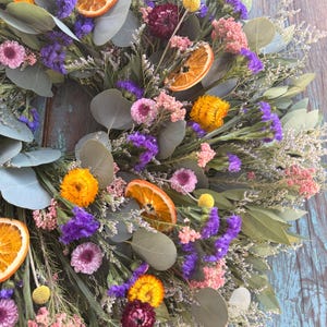 May include: A circular wreath composed of dried eucalyptus leaves, vibrant purple statice, pink flowers, yellow strawflowers, and dried orange slices. The arrangement features a mix of textures and colors, including deep burgundy accents and small yellow billy buttons.