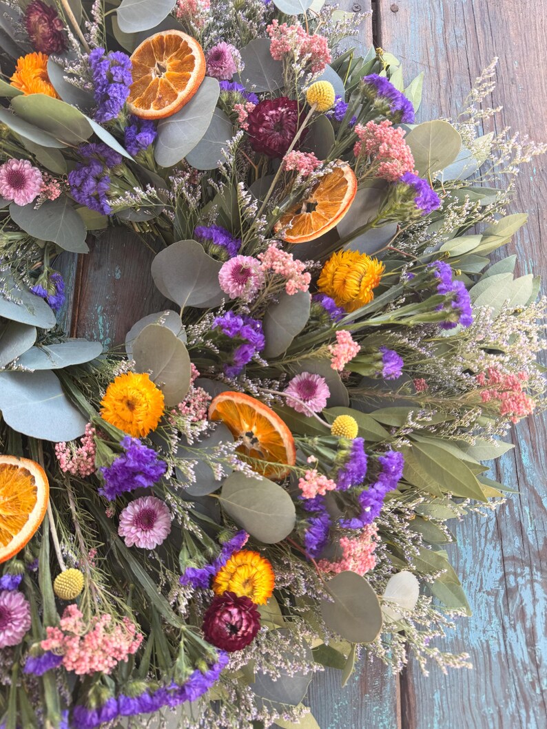 May include: A circular wreath composed of dried eucalyptus leaves, vibrant purple statice, pink flowers, yellow strawflowers, and dried orange slices. The arrangement features a mix of textures and colors, including deep burgundy accents and small yellow billy buttons.