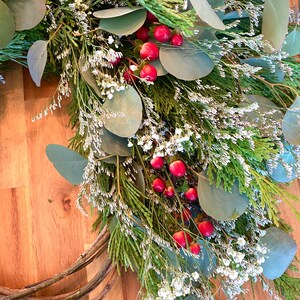 Op de afbeelding: Een handgemaakte krans met een mix van groen, waaronder eucalyptusbladeren, ceder takken en delicate witte bloemen. Heldere rode bessen zijn door de opstelling verspreid, wat een feestelijke en natuurlijke uitstraling cre&euml;ert. De krans is gemaakt op een natuurlijke wijnstokbasis.