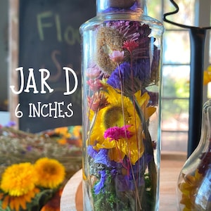 May include: A clear glass jar, approximately 15 cm tall, filled with dried flowers in shades of yellow, purple, pink, and green. The jar has a cork stopper. The text "JAR D" and "6 INCHES" are visible on the image.