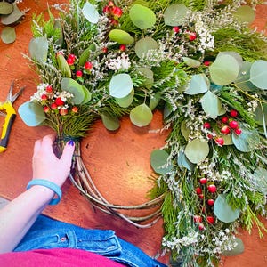 Op de afbeelding: Een gedeeltelijk voltooide krans met eucalyptusbladeren, kleine witte bloemen en rode bessen. De krans wordt geassembleerd op een houten oppervlak.
