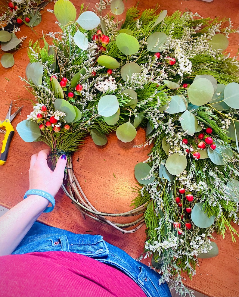 Op de afbeelding: Een gedeeltelijk voltooide krans met eucalyptusbladeren, kleine witte bloemen en rode bessen. De krans wordt geassembleerd op een houten oppervlak.