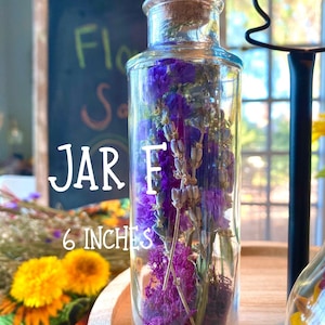 May include: A clear glass jar filled with purple dried flowers and moss, topped with a cork stopper. The jar is labeled "JAR E" and is 15 cm tall. The background includes a chalkboard and other floral arrangements.