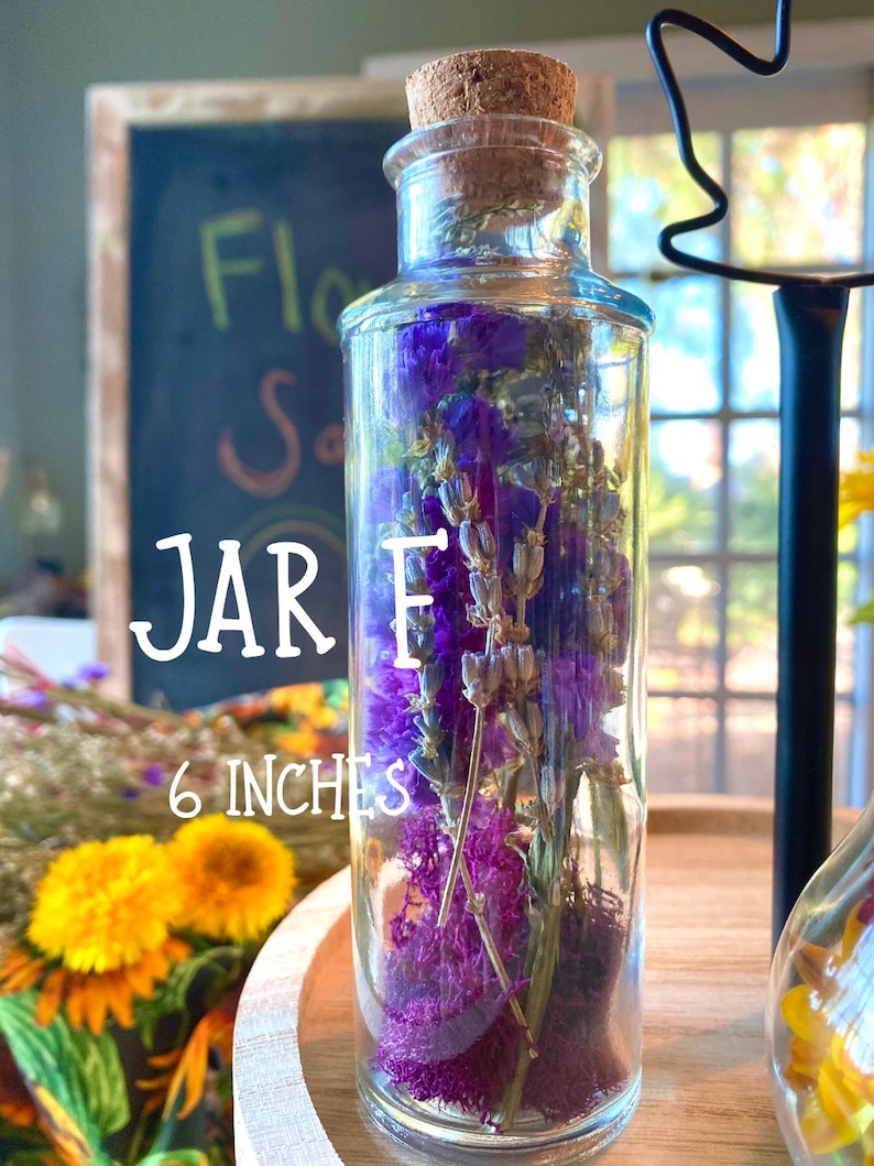 May include: A clear glass jar filled with purple dried flowers and moss, topped with a cork stopper. The jar is labeled "JAR E" and is 15 cm tall. The background includes a chalkboard and other floral arrangements.