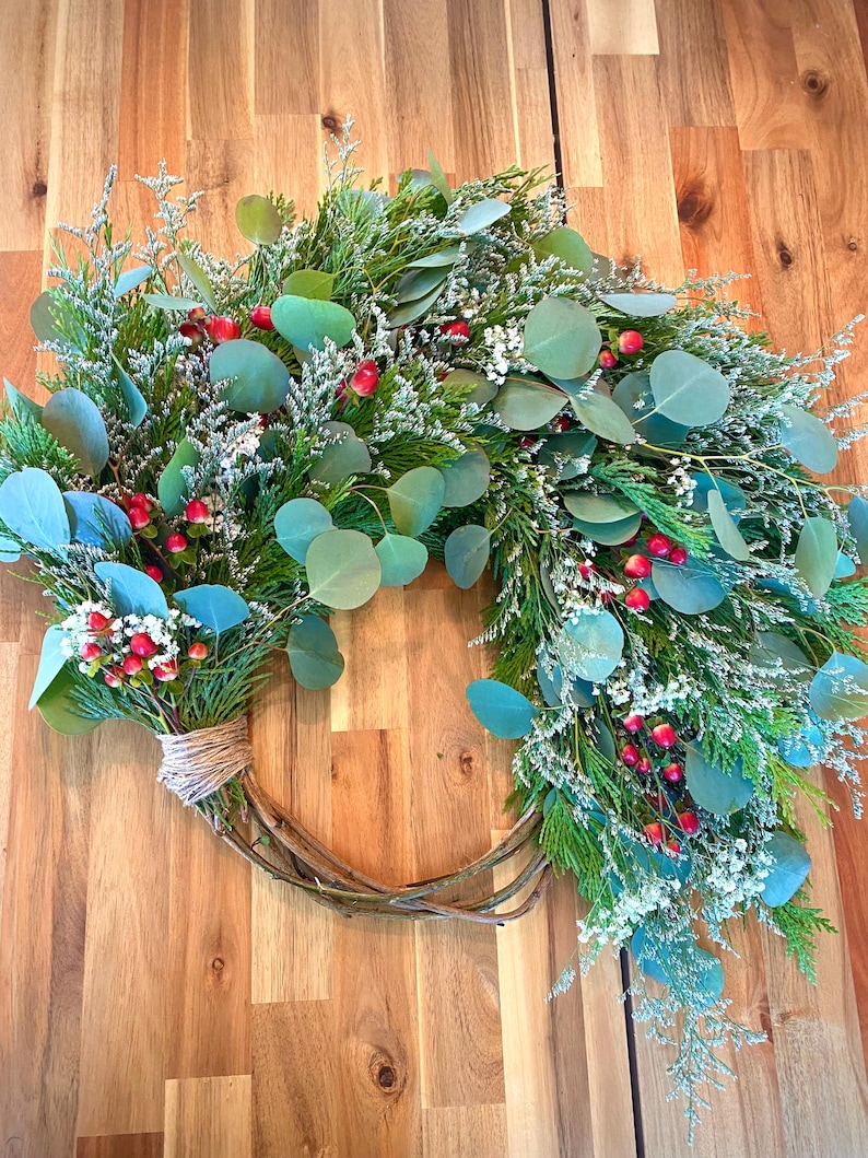 Op de afbeelding: Een handgemaakte krans met een basis van twijgen, versierd met groen gebladerte, eucalyptusbladeren, kleine witte bloemen en rode bessen. De krans is vastgebonden met touw, wat een natuurlijke, seizoensgebonden uitstraling geeft, geschikt voor huisdecoratie.