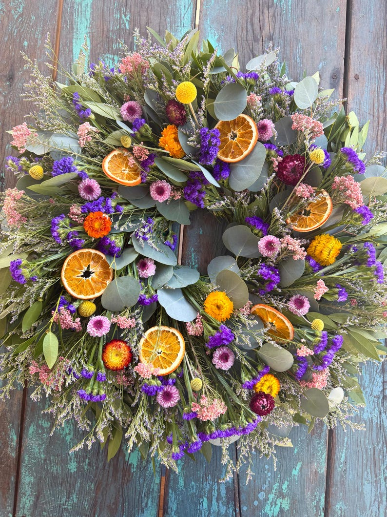 May include: A circular wreath featuring dried eucalyptus leaves, vibrant purple statice, pink filler flowers, yellow billy buttons, and dried orange slices. The arrangement is densely packed with various botanical elements, creating a textured, multi-colored display of preserved natural foliage and fruit.