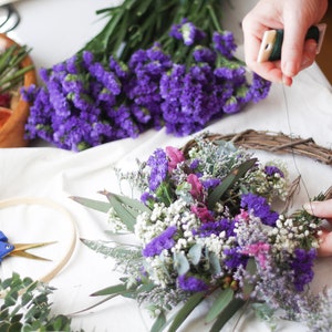 May include: A grapevine wreath base being decorated with purple statice, white baby's breath, and green eucalyptus leaves. Nearby are blue-handled floral shears, a wooden embroidery hoop, and a wooden bowl containing dried citrus slices.
