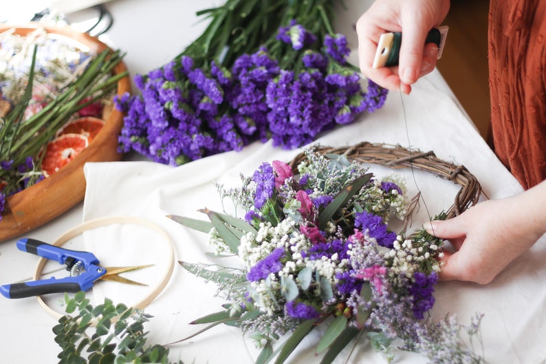 May include: A grapevine wreath base being decorated with purple statice, white baby's breath, and green eucalyptus leaves. Nearby are blue-handled floral shears, a wooden embroidery hoop, and a wooden bowl containing dried citrus slices.