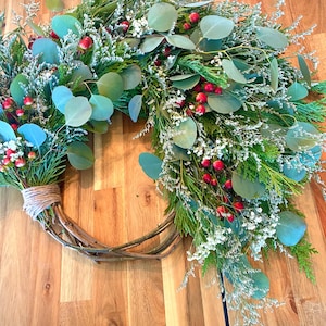 Op de afbeelding: Een handgemaakte krans met een natuurlijke wijnstokbasis, versierd met groene eucalyptusbladeren, witte bloemen, rode bessen en groenblijvende takjes. De krans is vastgebonden met touw, wat een feestelijke en seizoensgebonden decoratie cre&euml;ert.