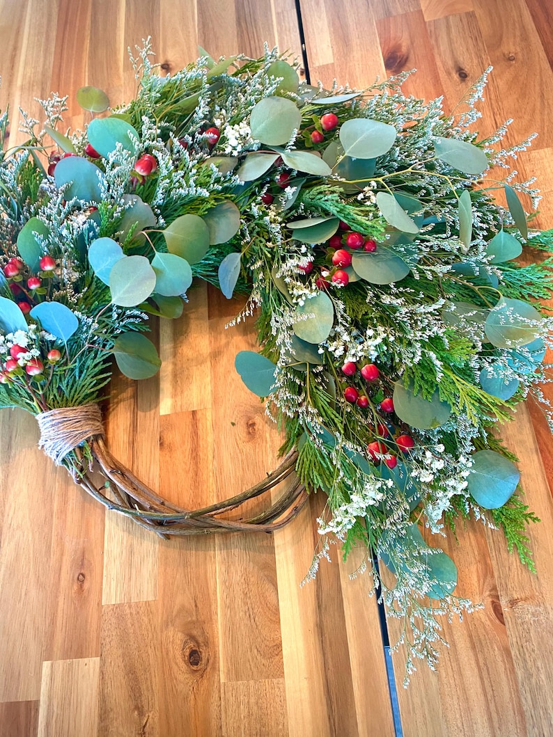 Op de afbeelding: Een handgemaakte krans met een natuurlijke wijnstokbasis, versierd met groene eucalyptusbladeren, witte bloemen, rode bessen en groenblijvende takjes. De krans is vastgebonden met touw, wat een feestelijke en seizoensgebonden decoratie cre&euml;ert.