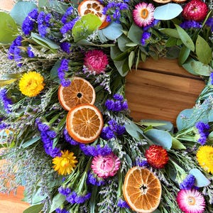 May include: A circular wreath composed of dried eucalyptus leaves, vibrant purple statice flowers, and bright yellow strawflowers. The arrangement is accented with pink strawflowers and several dried orange slices distributed throughout the foliage.