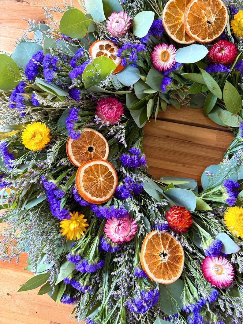May include: A circular wreath composed of dried eucalyptus leaves, vibrant purple statice flowers, and bright yellow strawflowers. The arrangement is accented with pink strawflowers and several dried orange slices distributed throughout the foliage.