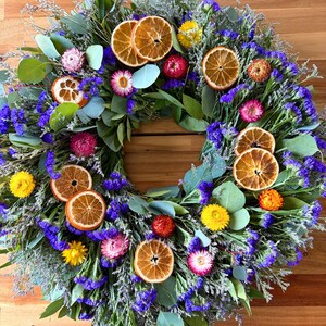 May include: A circular wreath featuring dried orange slices, vibrant purple statice flowers, green eucalyptus leaves, and small yellow, pink, and orange strawflowers. The arrangement is densely packed with various botanical elements.