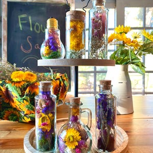 May include: Glass bottles filled with dried flowers are displayed on a two-tiered wooden tray. The bottles have cork stoppers. The flowers are yellow, purple, and pink. A chalkboard in the background reads "Flower Sale".