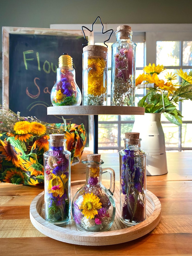 May include: Glass bottles filled with dried flowers are displayed on a two-tiered wooden tray. The bottles have cork stoppers. The flowers are yellow, purple, and pink. A chalkboard in the background reads "Flower Sale".