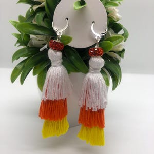 May include: A pair of dangle earrings with a white, orange, and yellow tassel design. The earrings have a silver hoop and a red crystal bead at the top.
