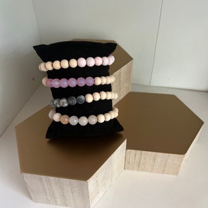 May include: Four beaded bracelets displayed on a black velvet stand. The bracelets feature a variety of beads, including light pink, lavender, gray, and natural wood tones. The bracelets are presented on a gold and white hexagonal display.