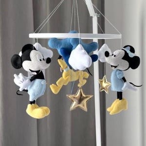 Puede incluir: Móvil para bebé blanco con figuras de peluche de Mickey y Minnie Mouse en azul y negro, con zapatos amarillos. El móvil también incluye adornos de estrellas doradas y una nube azul. El móvil cuelga sobre una cuna blanca.
