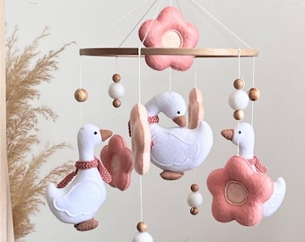 May include: A hanging mobile featuring three white felt geese with brown beaks and pink knitted scarves, interspersed with pink felt flowers and white and natural wood beads suspended from a circular wooden frame by white strings.