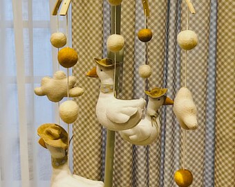 Op de afbeelding: Een baby mobile met witte ganzen met hoeden, vilten wolken en verschillende maten vilten ballen in crème en mosterdgeel. De mobiel hangt aan een houten boog en een centrale paal, ontworpen voor een kinderkamer.