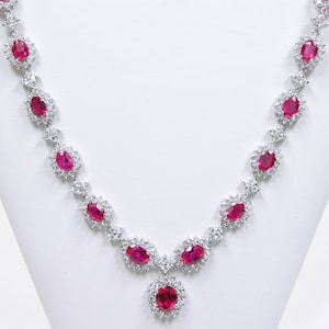 May include: A silver necklace with a pendant featuring a large oval ruby surrounded by smaller diamonds. The necklace is adorned with a row of oval rubies and diamonds.