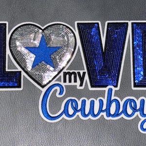 May include: A decorative patch featuring the text LOVE MY Cowboys with a silver sequin heart and a blue star. The letters L, V, E are filled with blue sequins. A ruler is visible on the left, showing the patch is approximately 5 inches wide.