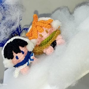May include: Two small crocheted figures with peach-colored skin. One figure has black hair, a white hood, and a blue scarf. The other has orange hair and a green sash. The text @hookedbyemi is visible in the bottom left corner of the image.
